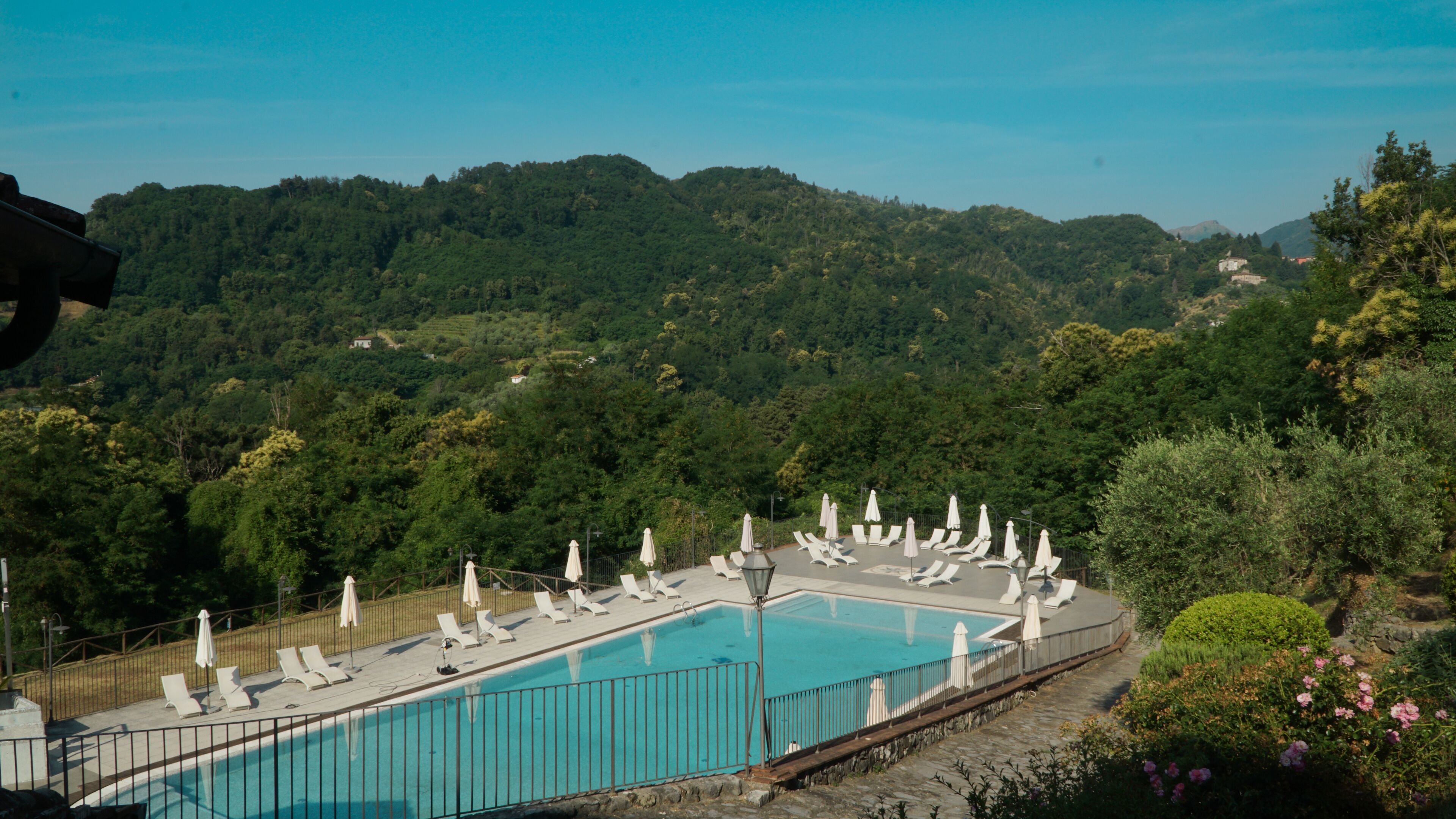 A rectangular pool with white lounge chairs and umbrellas, surrounded by a stone wall, set against a backdrop of lush green hills and a clear sky.