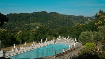 Ein rechteckiger Pool mit weißen Liegestühlen und Sonnenschirmen, umgeben von einer Steinmauer, vor einer Kulisse aus saftig grünen Hügeln und blauem Himmel.