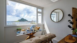 A room with a large window overlooking a coastal view, a glass-top table with various items, a round mirror, and a wooden wine rack.