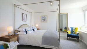 A four-poster bed with white linens, a bedside table with a lamp, and a chair with a blue and yellow cushion.