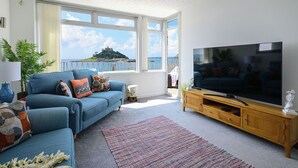 A living room with a large TV, blue sofas, a wooden TV stand, and a view of the sea.