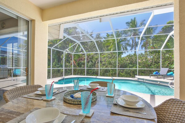An indoor pool area with clear glass walls, a dining table set with bowls and glasses, and a view of palm trees outside.