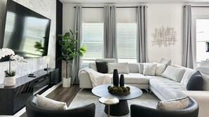 Un salon moderne avec un canapé d’angle, un téléviseur à écran plat, un ventilateur de plafond et une plante décorative.