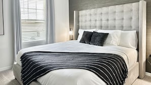 Une chambre à coucher avec une tête de lit capitonnée, un lit avec des oreillers blancs et foncés, un plaid à rayures et un ventilateur de plafond.