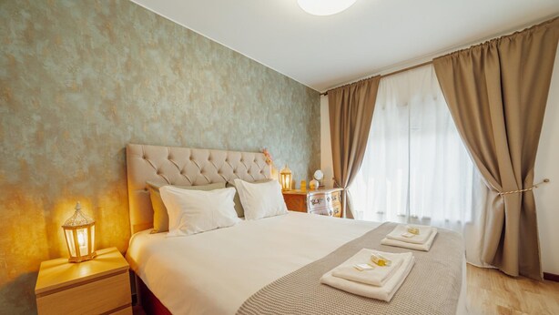 Habitación de hotel con una cama grande, una mesita de noche con lámpara, una mesita auxiliar y una ventana con cortinas.