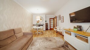 Sala de estar con sofá, mesa de comedor con sillas, televisor montado en la pared y área de cocina con piso de madera.