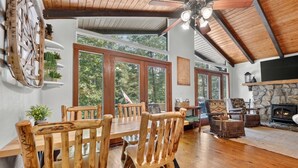 A wooden dining table with chairs, a stone fireplace, and large windows with a view of trees. - At the End of the Road. Toccoa River Front (Fannin County)