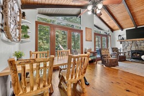 Ein Holzespeisetisch mit Stühlen, ein Steinkamin und große Fenster mit Blick auf Bäume.
