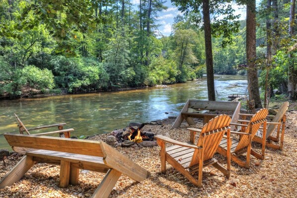 A riverside campsite with a fire pit and wooden chairs.