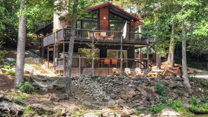 A cabin with a stone foundation, wooden deck, and a red roof, surrounded by trees and a body of water. - At the End of the Road. Toccoa River Front (Fannin County)