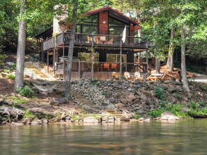 Eine Holzhütte mit Stein Fundament, einer Holzterrasse und einem roten Dach, umgeben von Bäumen und einem Gewässer.