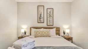 Ein ordentlich bezogenes Bett mit weißer Decke und grauer Strickdecke, neben dem sich zwei Nachttische mit Lampen und dekorativen Vasen befinden.