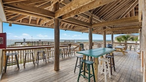 Terraza de madera con mesas y taburetes, con vista a una playa.