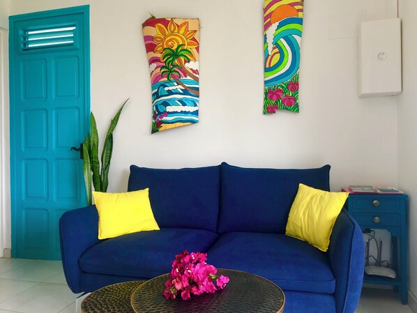 Une pièce avec un canapé bleu, deux coussins jaunes, une table basse en verre ornée de fleurs roses, une porte de couleur turquoise et deux décorations murales colorées.