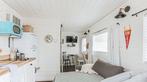 Une pièce compacte et bien éclairée, dotée d’une kitchenette, d’un lit recouvert d’une courtepointe, d’un téléviseur fixé au mur et d’une petite table avec des chaises.