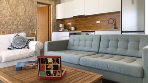 Un salon moderne avec un canapé gris, un fauteuil blanc et une table basse en bois. On aperçoit, au fond, une cuisine avec des meubles blancs et un plan de travail en bois.