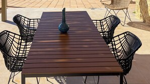Une terrasse extérieure avec une table en bois et des chaises en fil de fer noir, un mur en pierre et une vue sur la mer et les collines.