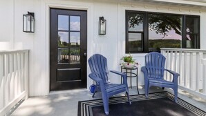 Eine Veranda mit zwei blauen Adirondack-Stühlen, einem kleinen runden Tisch mit einem Blumentopf und einer schwarzen Tür mit einem Fenster.
