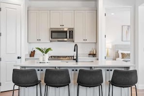 A modern kitchen with white cabinets, a stainless steel microwave, a gas stove, and a bar area with three black chairs.