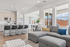 A modern living space with a grey sectional sofa, a dining area, and a kitchen with white cabinetry.