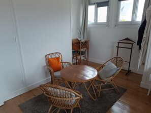 A room with a round wooden table, wicker chairs, a grey rug, and a window with curtains. - House with garden 30 metres from the beach (Ver-sur-Mer)