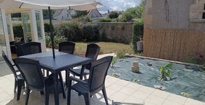 An outdoor patio with a table and chairs, a covered seating area, and a garden with a water feature. - House with garden 30 metres from the beach (Ver-sur-Mer)