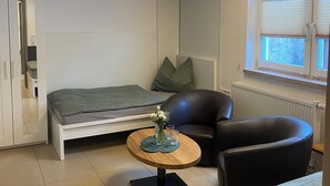 A compact, modern room with a bed, a small round table with a vase and glasses, a black leather chair, and a window with blinds.