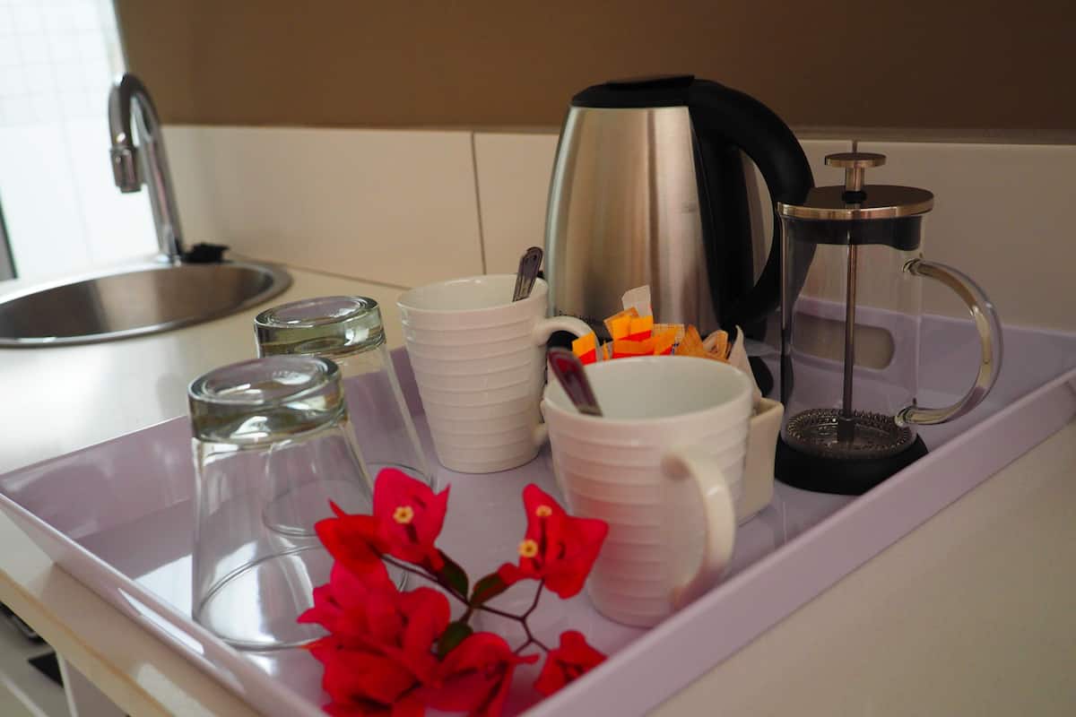 Una encimera de cocina con una cafetera, un hervidor, una pila de tazas blancas, un vaso y una bandeja con flores rojas.