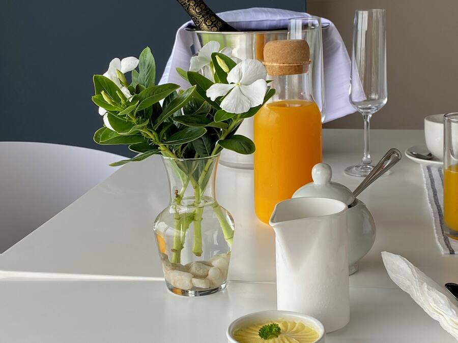 Una mesa con un florero de vidrio transparente con flores blancas, una botella alta con tapón de corcho, una jarra blanca, un tazón pequeño con decoración y un vaso con pajita.