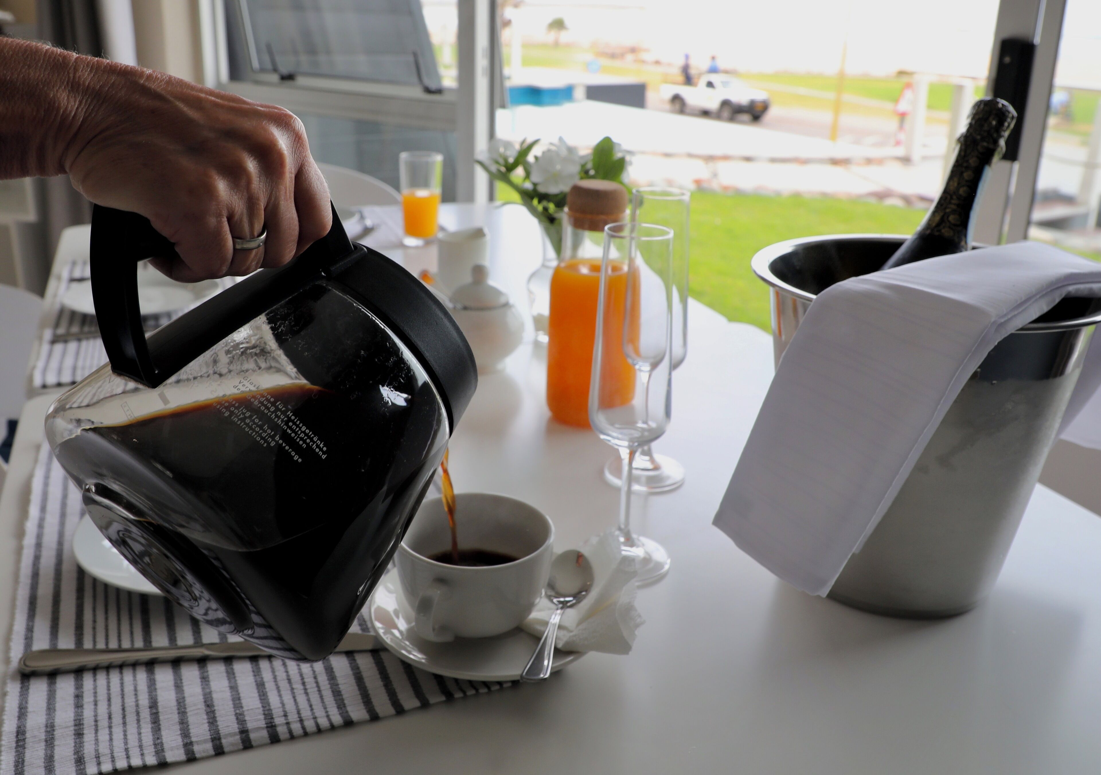 Una mano sirve café en una taza sobre una mesa, con vista a un espacio verde y un vehículo afuera.