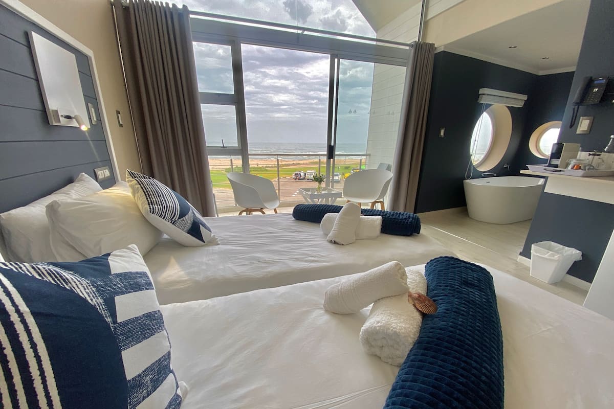 Una habitación de hotel moderna con dos camas, un ventanal con vista a la playa, un baño con bañera y una zona de estar con una silla y una mesa.