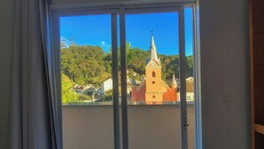 Une pièce dotée d’une porte-fenêtre coulissante donnant sur un balcon. Le balcon offre une vue sur une église avec un clocher et sur la végétation environnante.