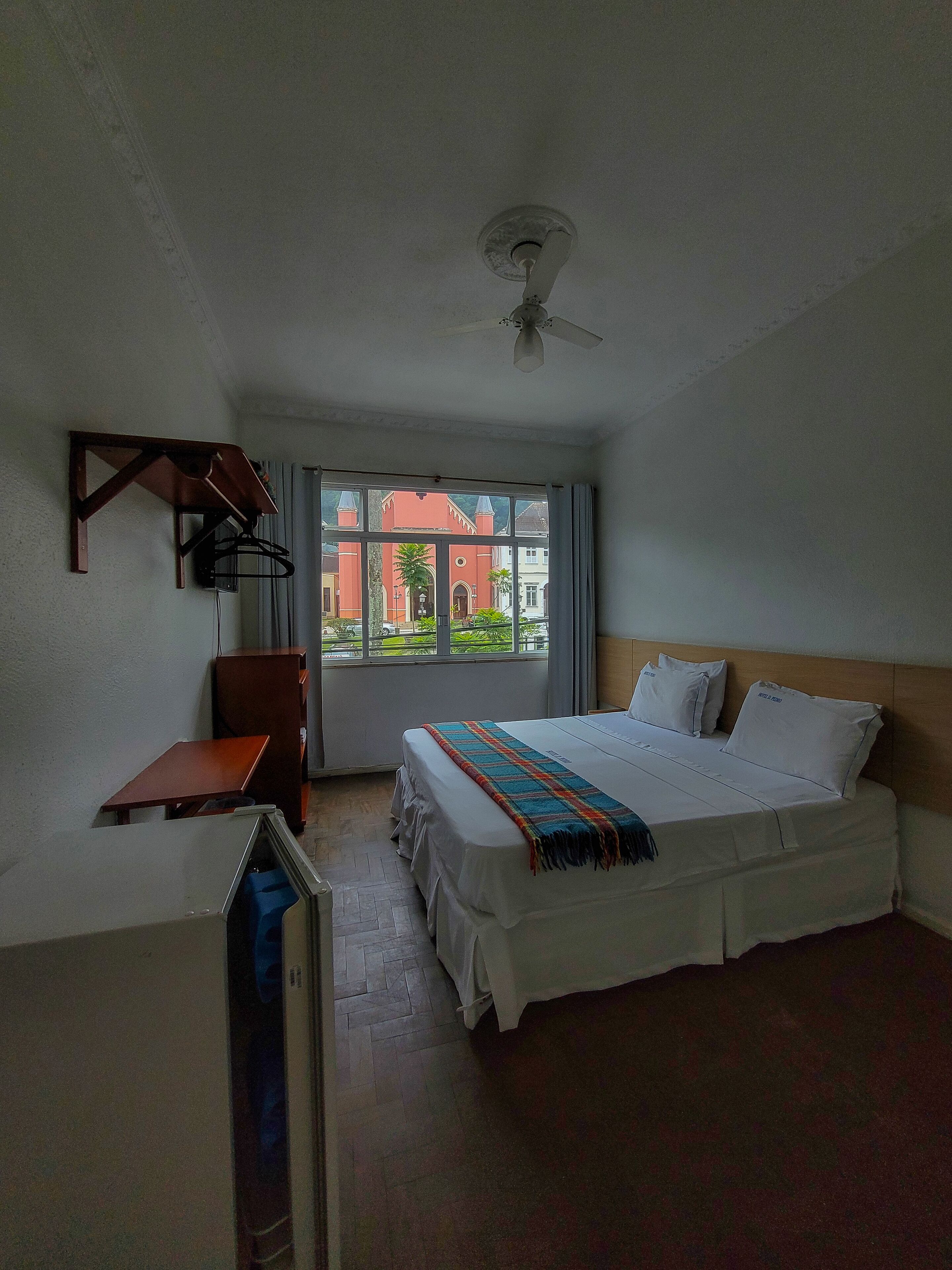 Quarto de hotel com cama, escrivaninha, ventoinha no teto e janela com vista para prédios.