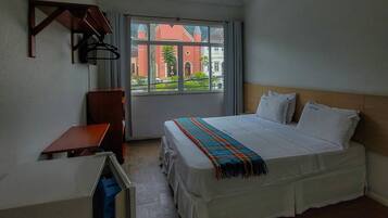 Quarto de hotel com cama, escrivaninha, ventoinha no teto e janela com vista para prédios.