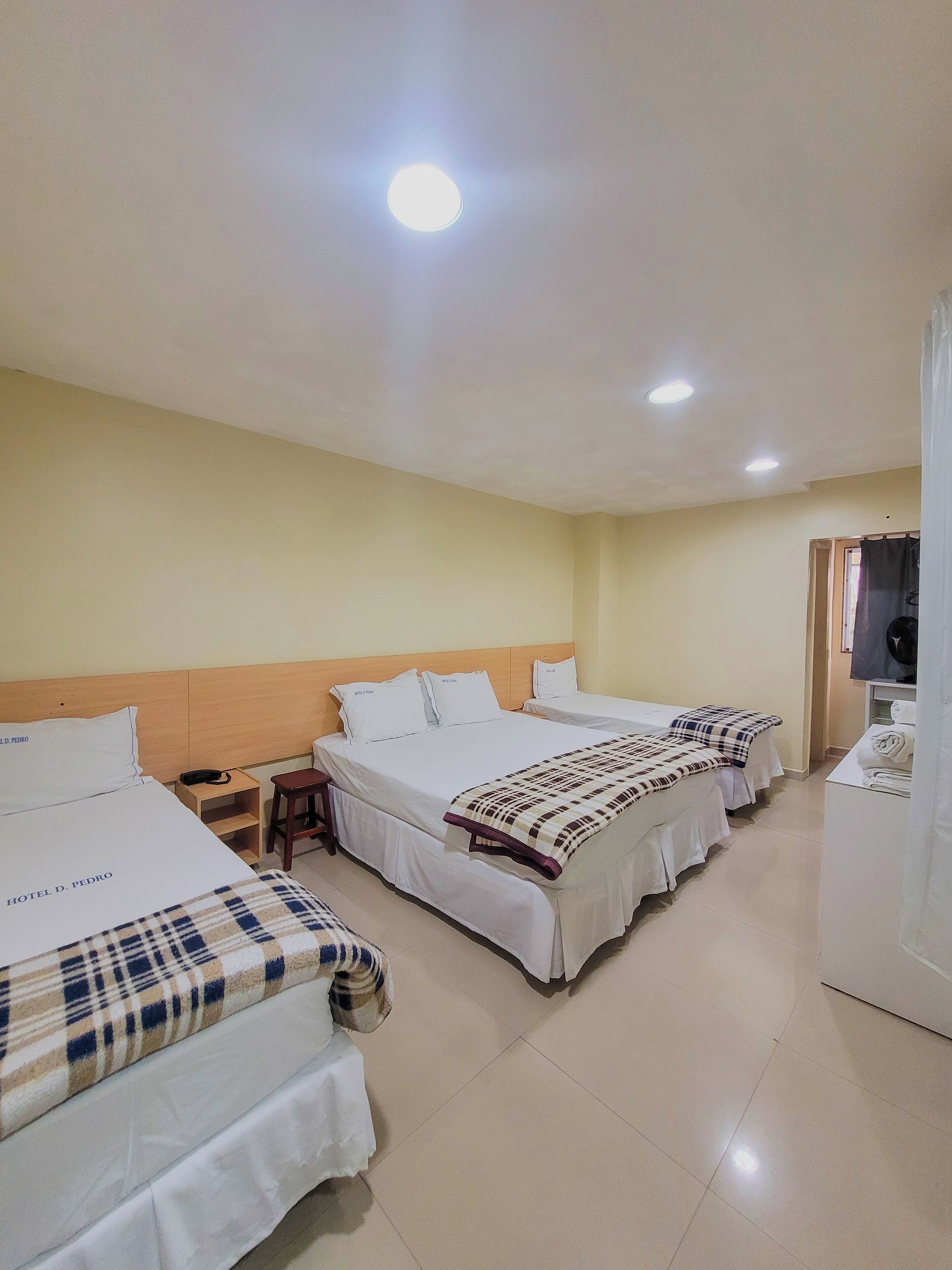 Habitación de hotel con tres camas, un cabecero de madera, una mesita de noche pequeña con una lámpara y un televisor sobre un soporte.