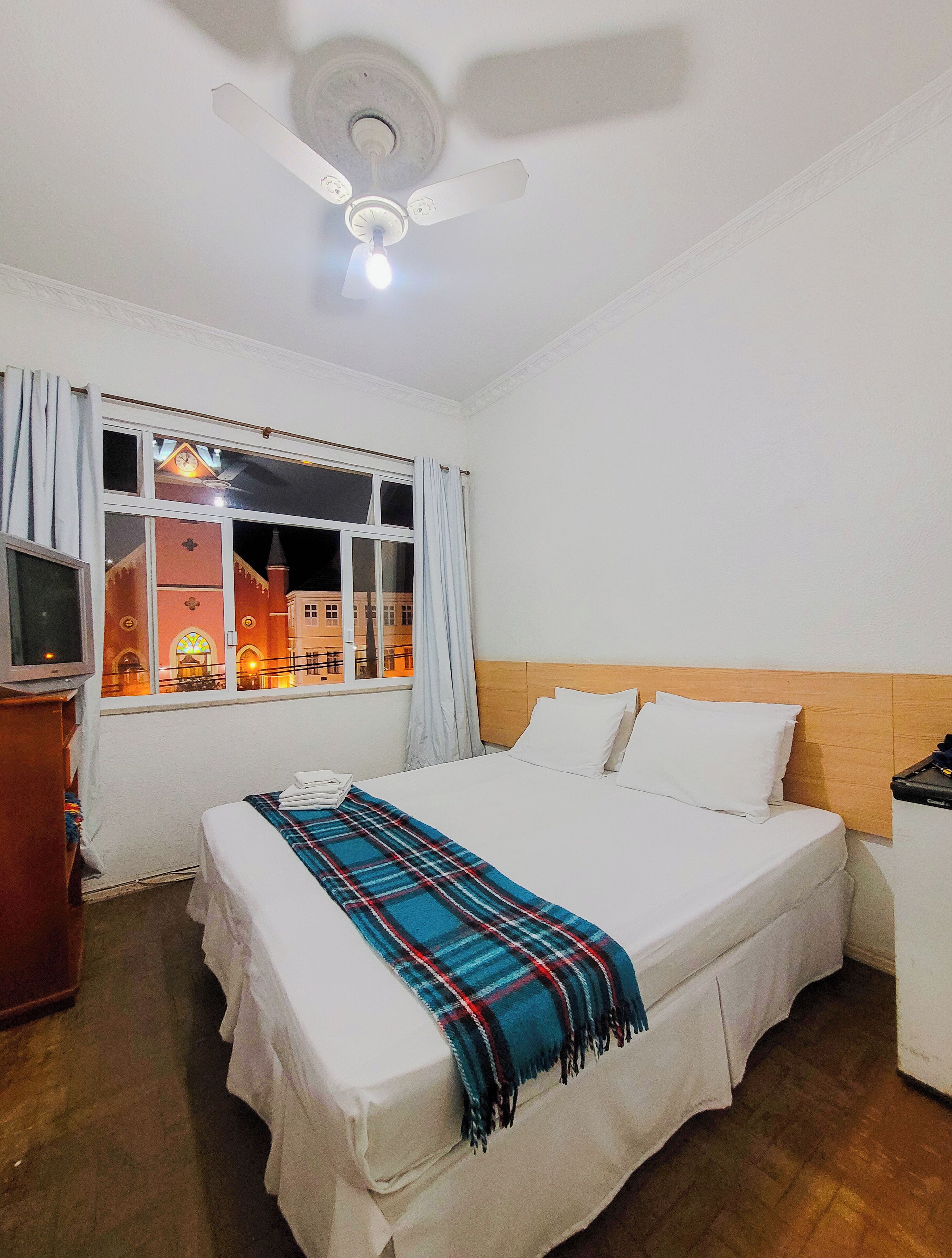 Une chambre d’hôtel équipée d’un lit, d’une télévision, d’un ventilateur de plafond et d’une fenêtre avec des rideaux.