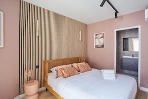 A bedroom with a bed, a bedside lamp, a wall-mounted light, and a bathroom with a mirror and sink.