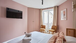Un cuarto de hotel con una cama, un televisor montado en la pared, dos sillas, un ventanal con cortinas y un cuadro enmarcado.