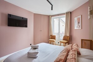 A hotel room with a bed, a TV mounted on the wall, two chairs, a window with curtains, and a framed picture on the wall.