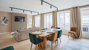 Una sala de estar moderna con un sofá, una mesa de comedor y sillas. Hay un televisor montado en la pared y ventanas amplias con cortinas.