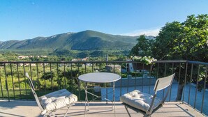 Un balcón con una mesa y dos sillas, con vistas a un paisaje montañoso con vegetación y edificios a lo lejos.