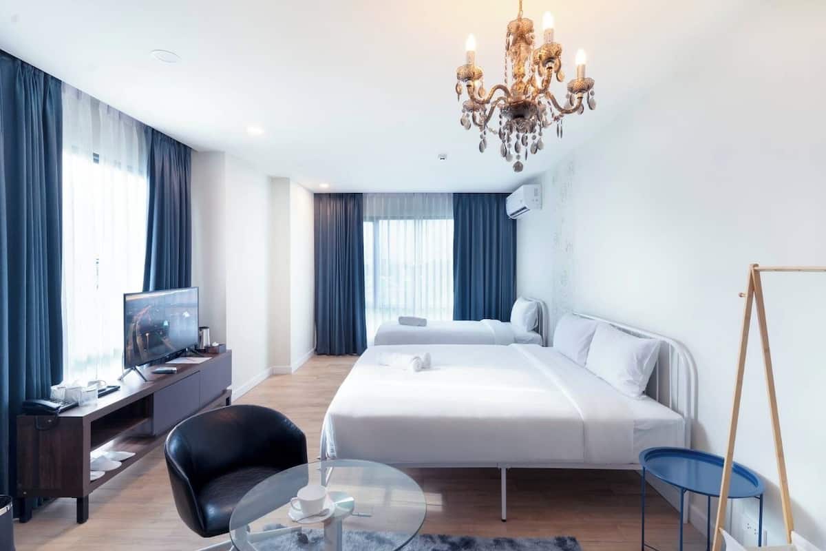 A modern hotel room with a large bed, a desk with a computer, a chair, a small table, and a chandelier.