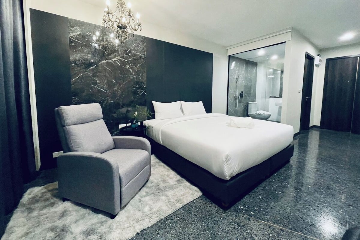 A modern hotel room with a large bed, a grey armchair, a chandelier, and a marble wall.