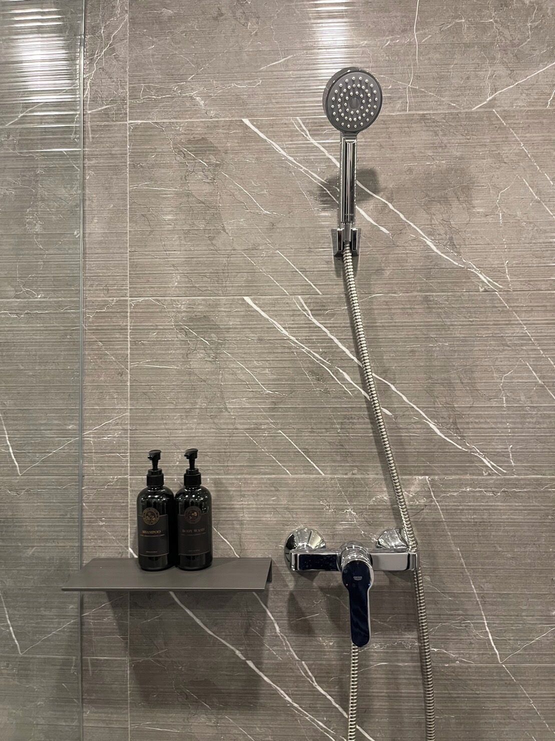 Een badkamer met een douche, een handdouche, en twee flessen shampoo op een plank.