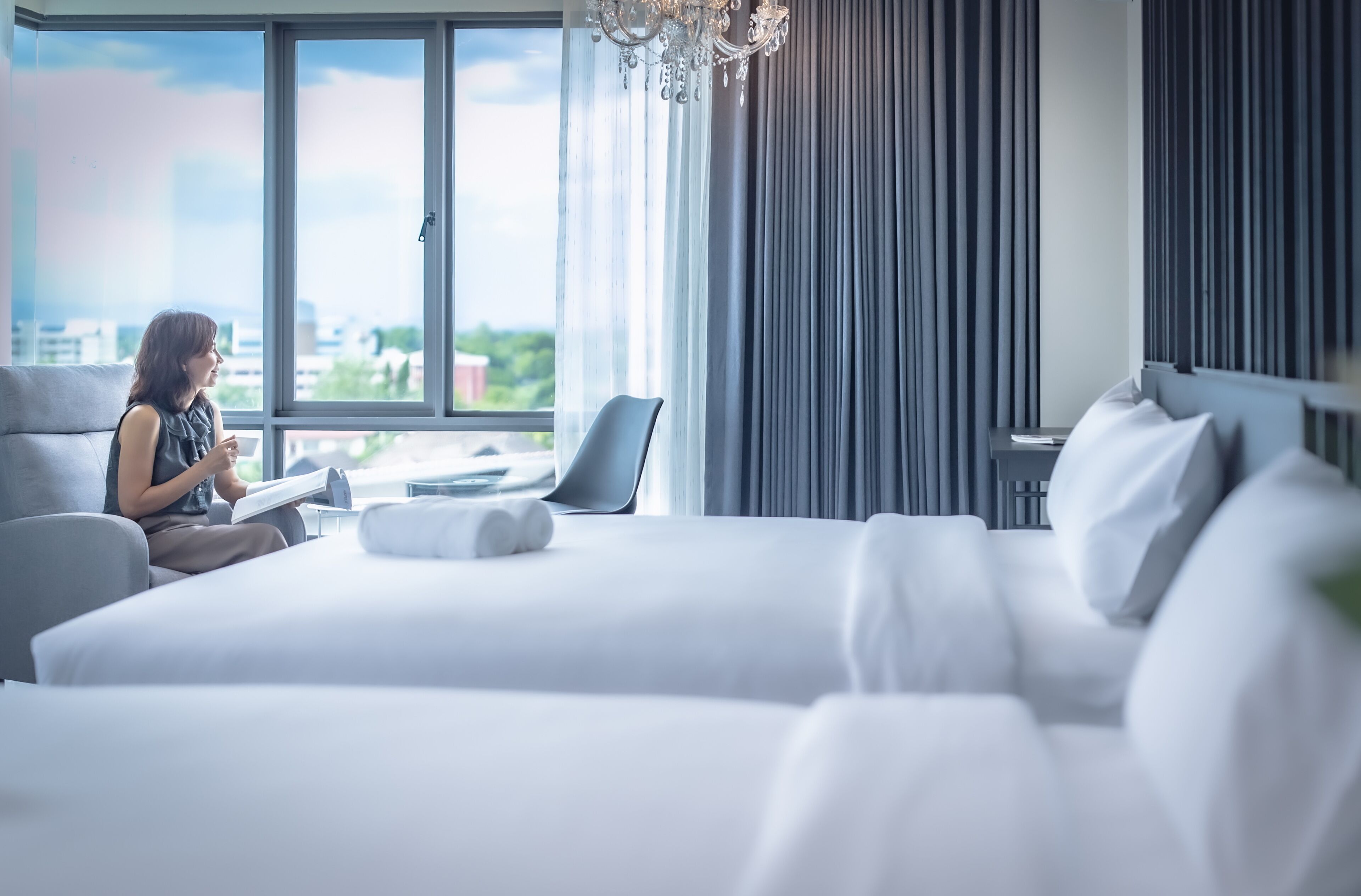 Une chambre d’hôtel moderne dotée d’un grand lit, d’un coin salon avec un fauteuil et offrant une vue sur la ville grâce à de grandes fenêtres.