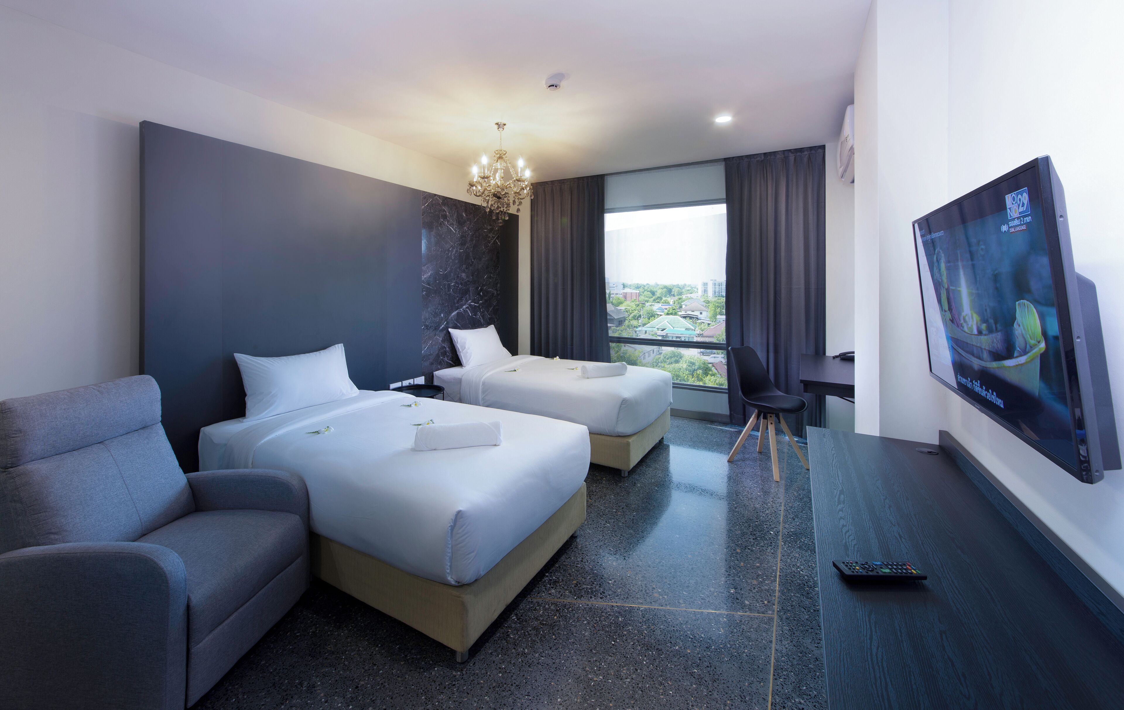 A hotel room with two beds, a grey armchair, a TV on the wall, and a view of the city through the window.