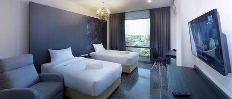 A hotel room with two beds, a grey armchair, a TV on the wall, and a view of the city through the window.