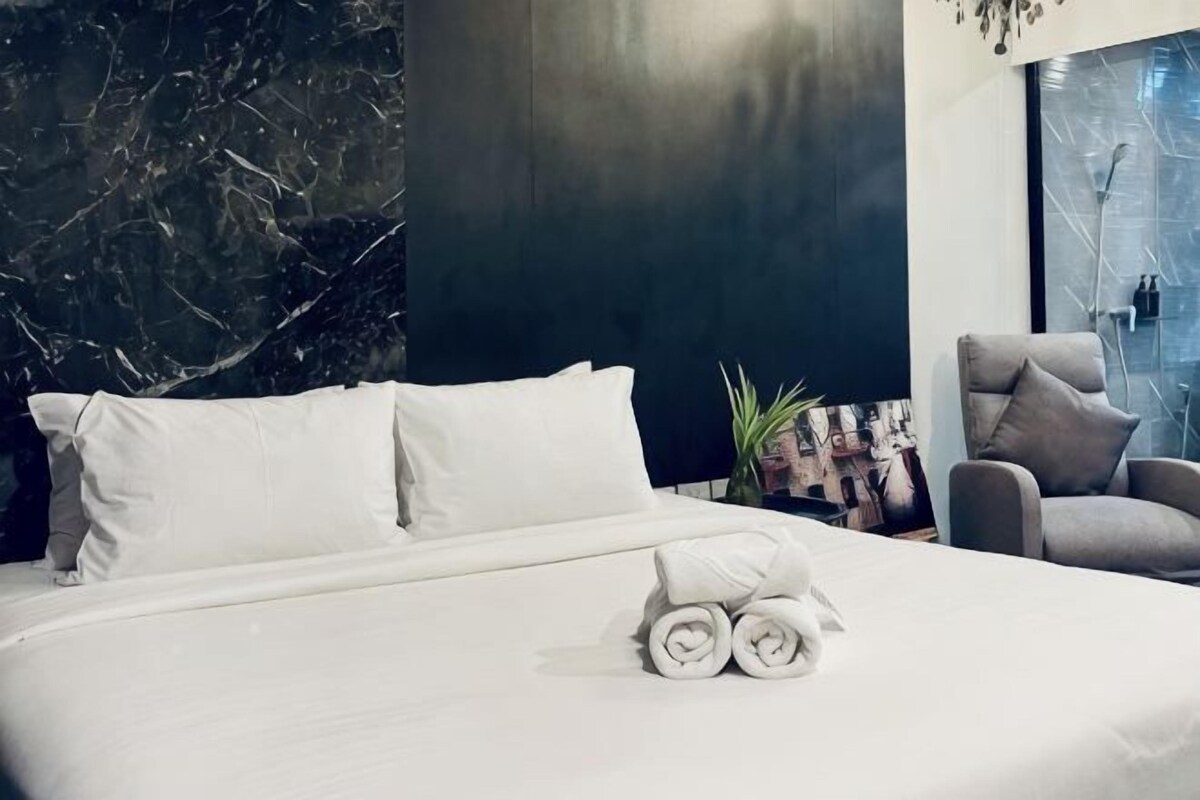A neatly made bed with white linens and rolled towels, a dark wall with marble-like texture, a grey armchair, and a glass door with a view.