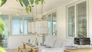 Un comedor moderno con una larga mesa de madera dispuesta para cuatro personas, asientos blancos acolchados e iluminación colgante.