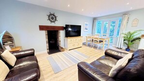 A living room with leather sofas, a TV, a fireplace, a dining table with chairs, and large windows. - Stunning Coastal Cottage - Sleeping 6 in 2 Bedrooms - Pool and Beach Access Included. (Lovely Central Location - Detached)